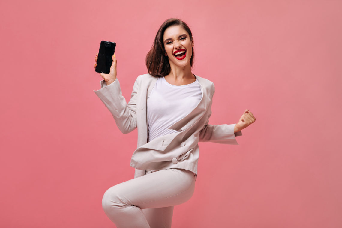 happy lady beige suit poses with phone pink background joyful girl office dress with red lips holds smartphone min